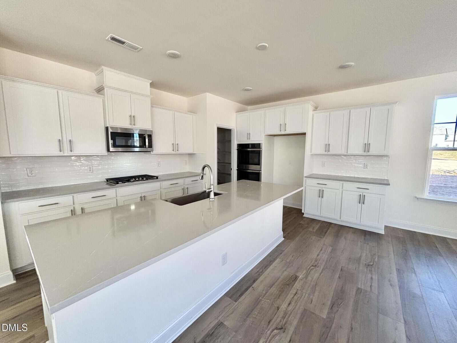 Modern kitchen with white cabinets, stainless steel appliances, an island with a sink, light gray countertops, tile backsplash, and wood-style flooring. Large windows provide natural light.