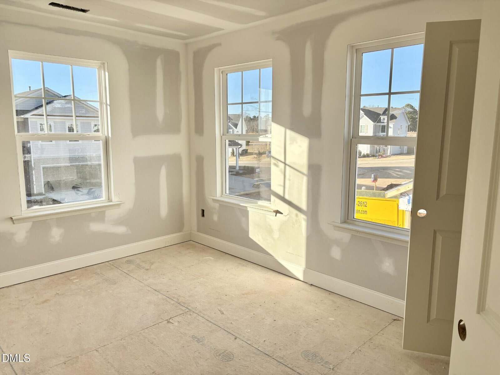 A bright, unfinished room with drywall installed and unpainted, featuring three large windows letting in sunlight, and a door slightly open. The floor is bare, and houses and a yellow dumpster are visible outside.