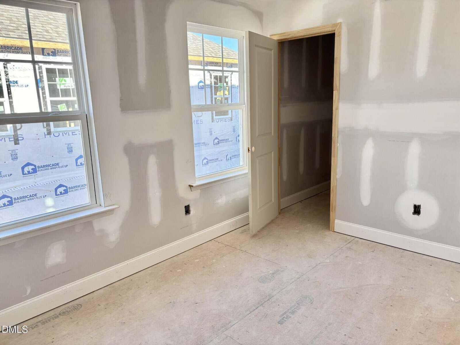 A room under construction with unfinished drywall, two windows, and a door leading to a small closet. The floor is bare, and there are visible wall seams and outlets.