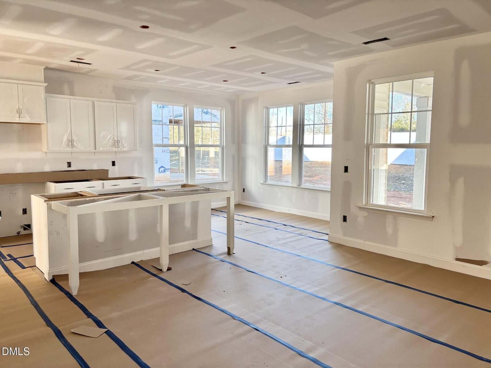 A bright, unfinished kitchen and dining area with white walls, unpainted drywall, and windows letting in sunlight. The floor is covered with protective paper and blue tape, and white cabinets are partially installed.