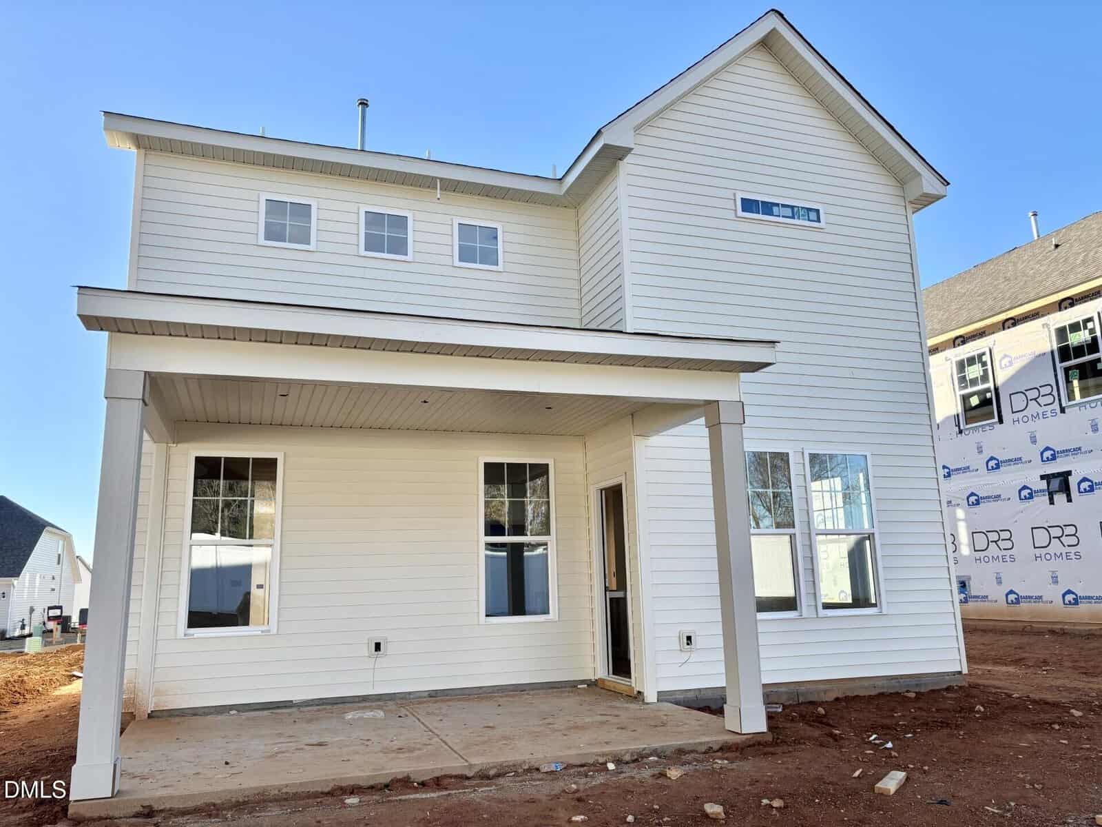 A newly constructed two-story house with white siding, large windows, and a covered front porch. The yard has bare dirt, and neighboring homes are visible, including one under construction.