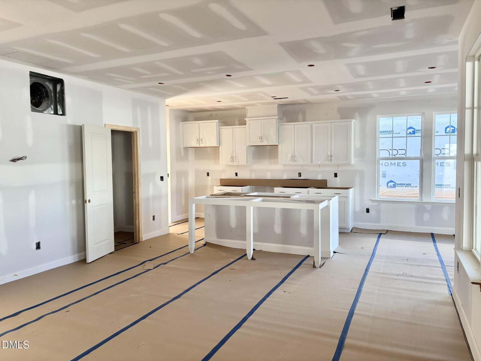 A newly constructed kitchen with white cabinets, an island, and large windows. The room is unfinished, with drywall seams visible and protective paper covering the floor.