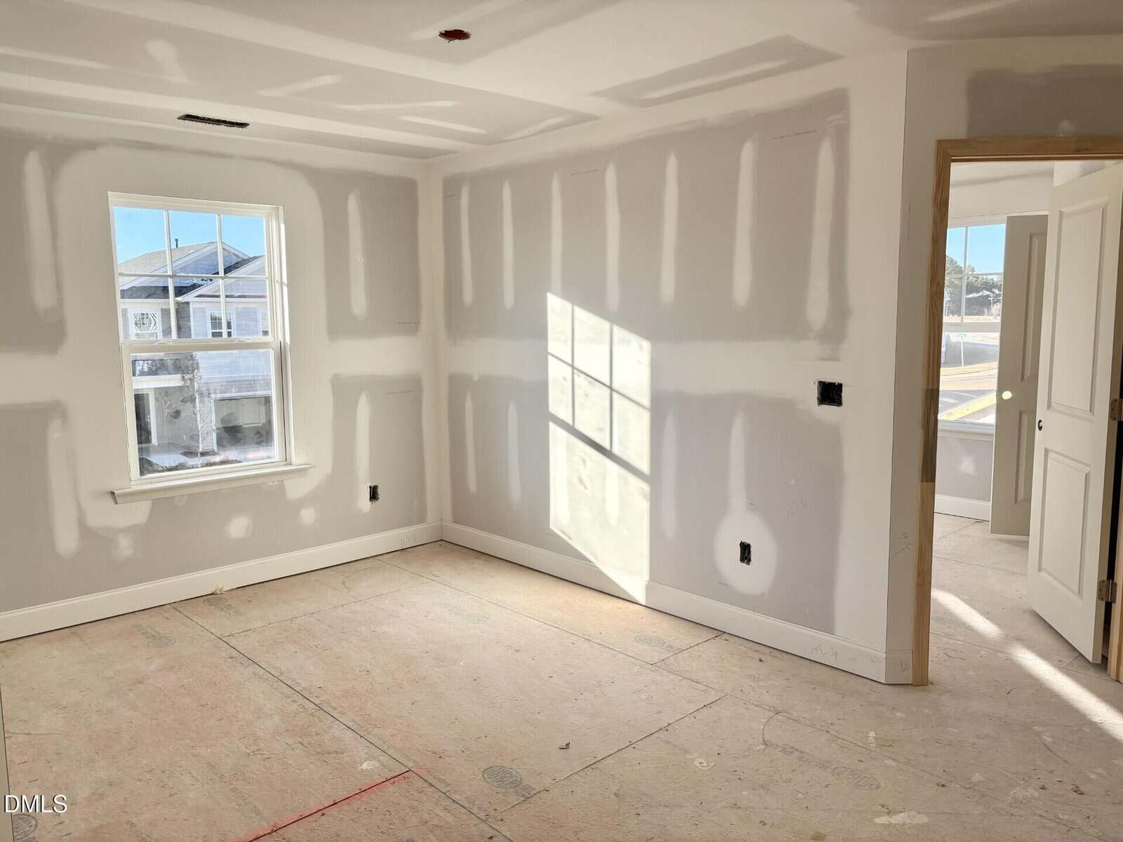 A bright, unfinished room with drywall panels and visible seams, a window letting in sunlight, an open door, and bare subflooring. No furniture or decorations are present.