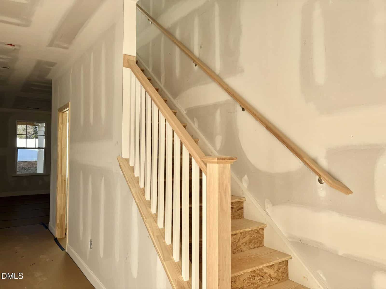 Unfinished wooden staircase with white balusters and a handrail, set against drywall that is partially primed for painting. Sunlight enters through a window at the end of the hallway.