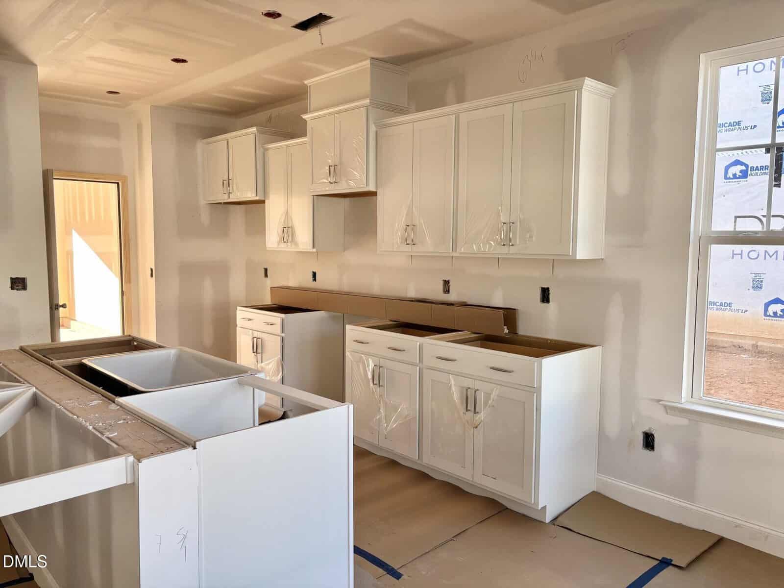 A kitchen under construction with white cabinets installed, some covered in protective plastic. Countertops are missing, and appliances are not yet in place. Walls are unfinished drywall, and there is a window letting in natural light.