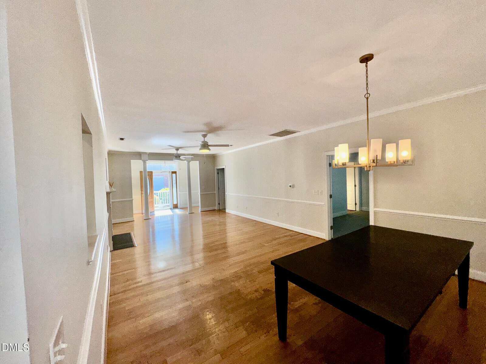 A spacious room with light-colored walls, wooden floors, a ceiling fan, a modern light fixture over a dark dining table, and large windows and doors letting in natural light at the far end.