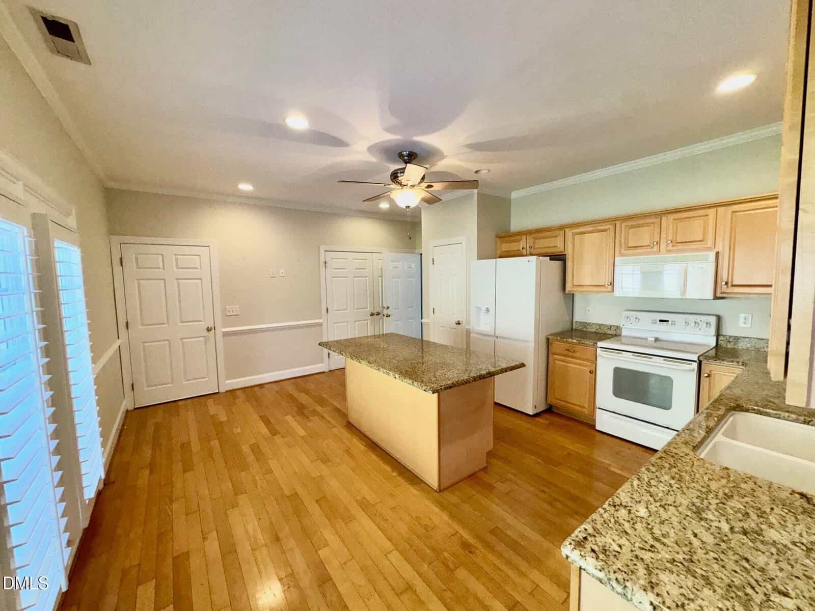 A spacious kitchen with wood floors, light wood cabinets, a central island with granite countertop, white appliances, sink, and multiple doors along the walls. Ceiling fan with lights is installed above the island.
