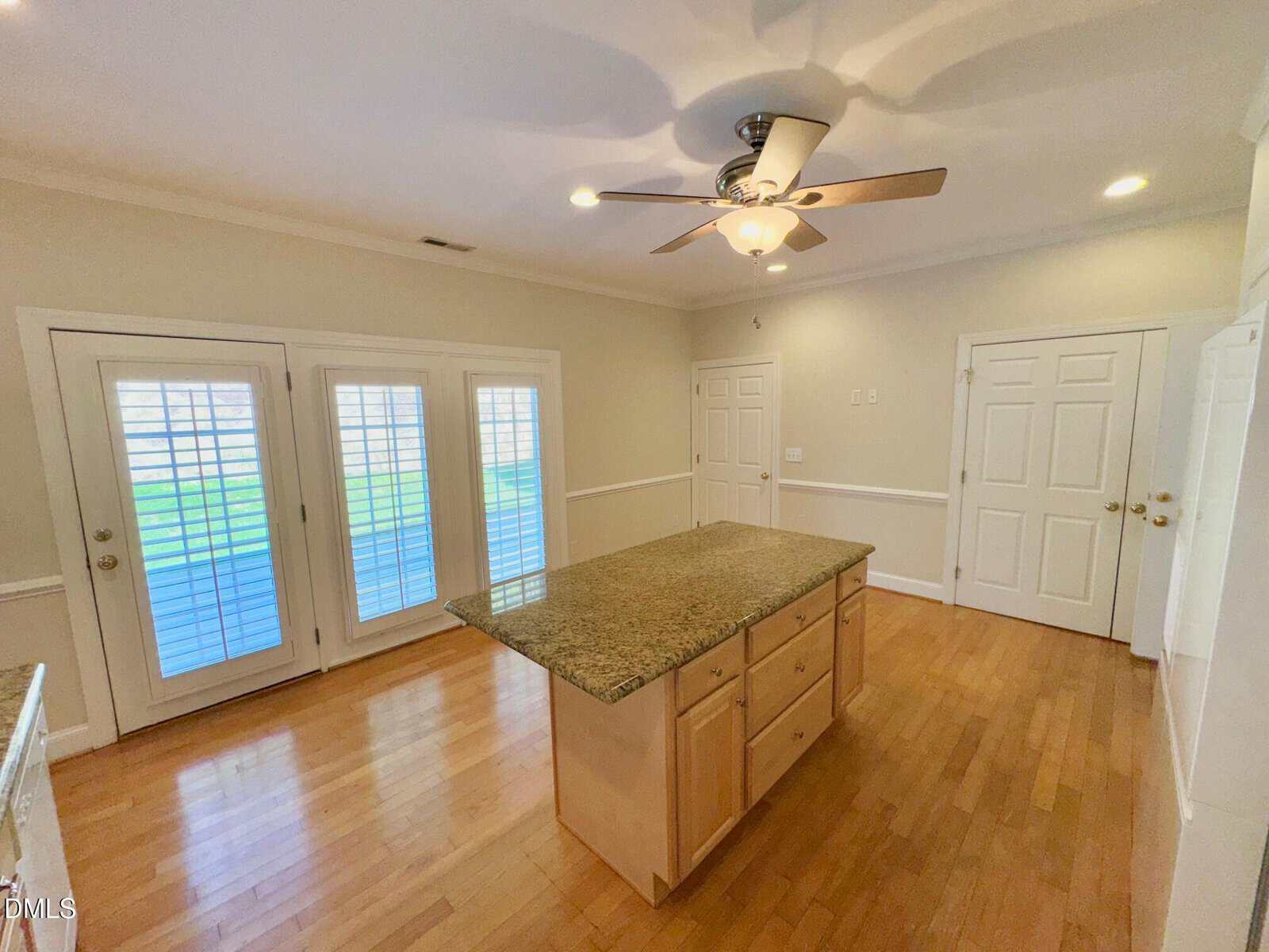 A kitchen with hardwood floors, a central island with a granite countertop, light wood cabinets, double doors with blinds, and several white doors on the walls. A ceiling fan with lights is above the island.