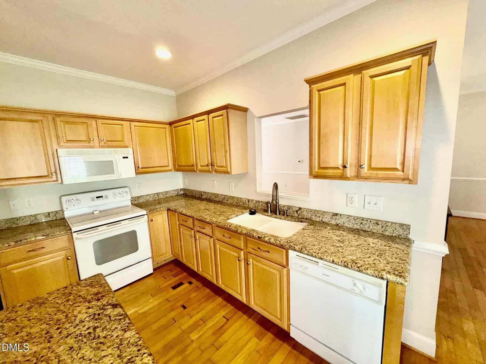 A kitchen with light wood cabinets, granite countertops, a white stove, microwave, dishwasher, and sink. The walls are light-colored, and the floor is wood. There is a cutout window above the sink.