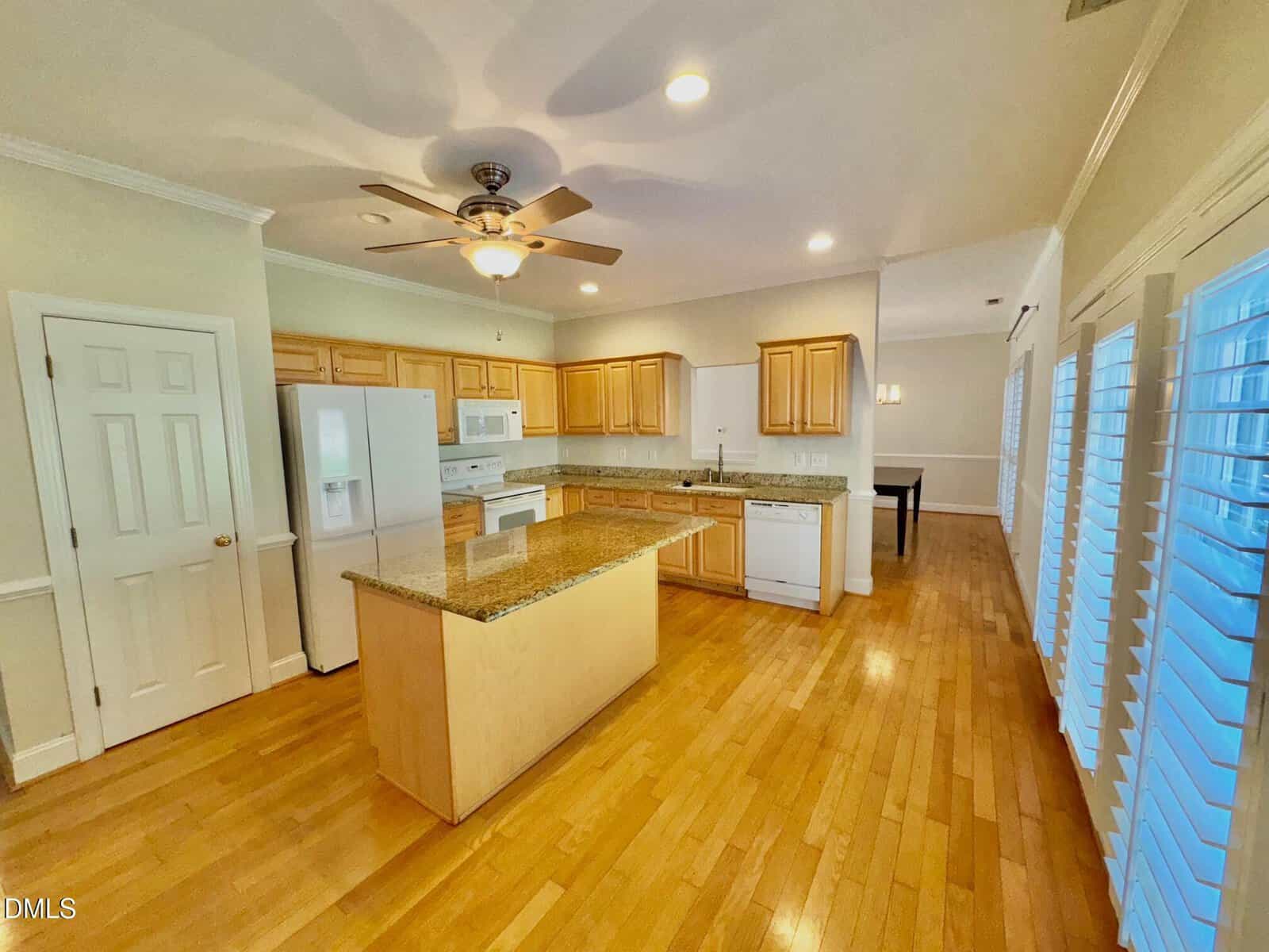 A spacious kitchen with light wood floors, light wood cabinets, granite countertops, a center island, and white appliances. Large windows with blinds line one wall, and a ceiling fan is mounted above the kitchen area.