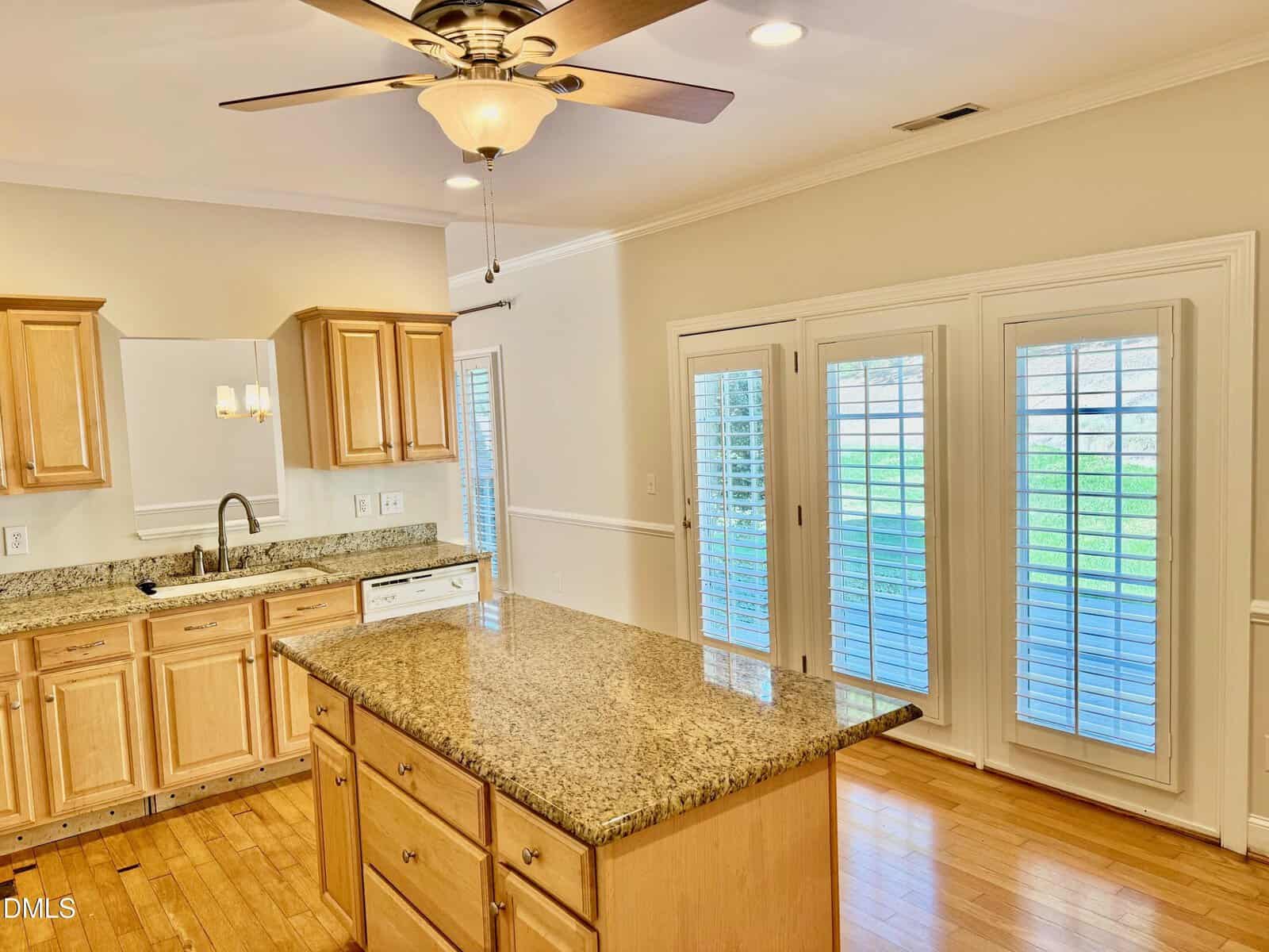 Bright kitchen with light wood cabinets, a granite island, stainless sink, and white appliances. French doors with white blinds lead outside. Ceiling fan with light fixture is above the island. Hardwood floors throughout.