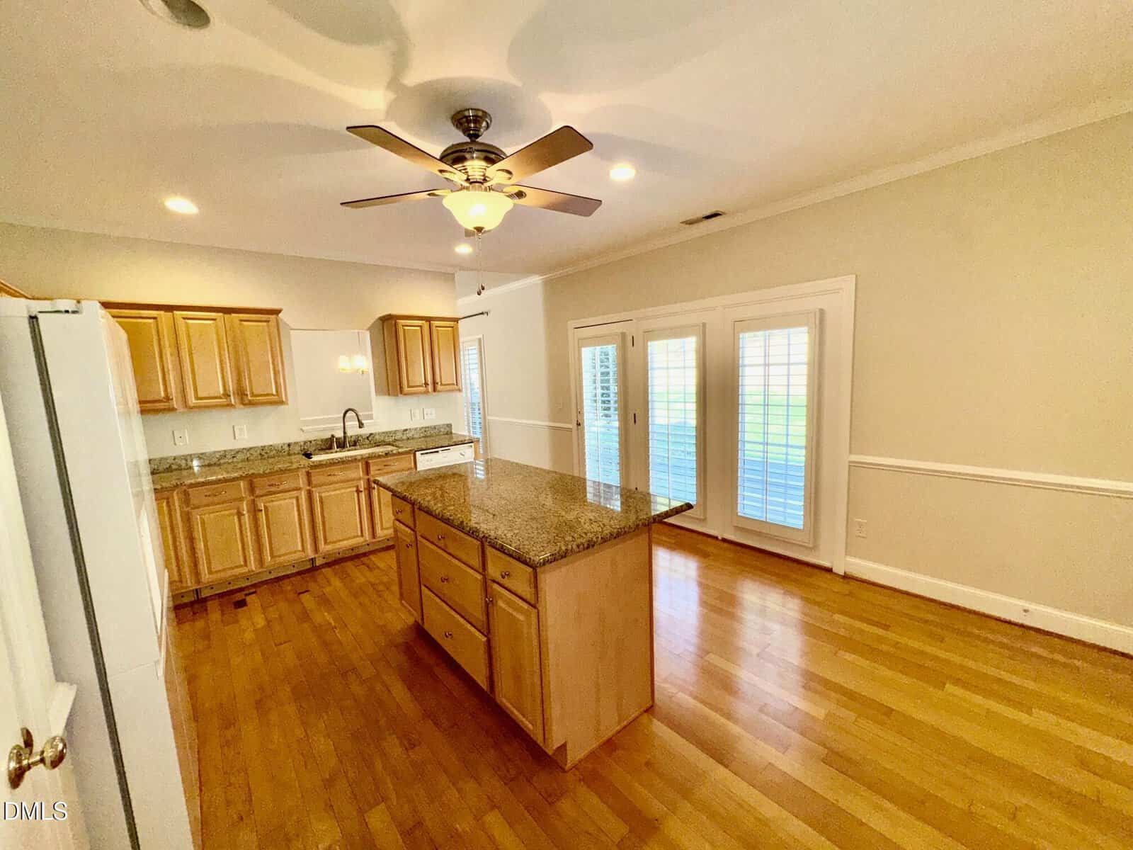 A kitchen with wooden cabinets, granite countertops, an island in the center, hardwood floors, a ceiling fan with lights, and French doors with white blinds leading outside.