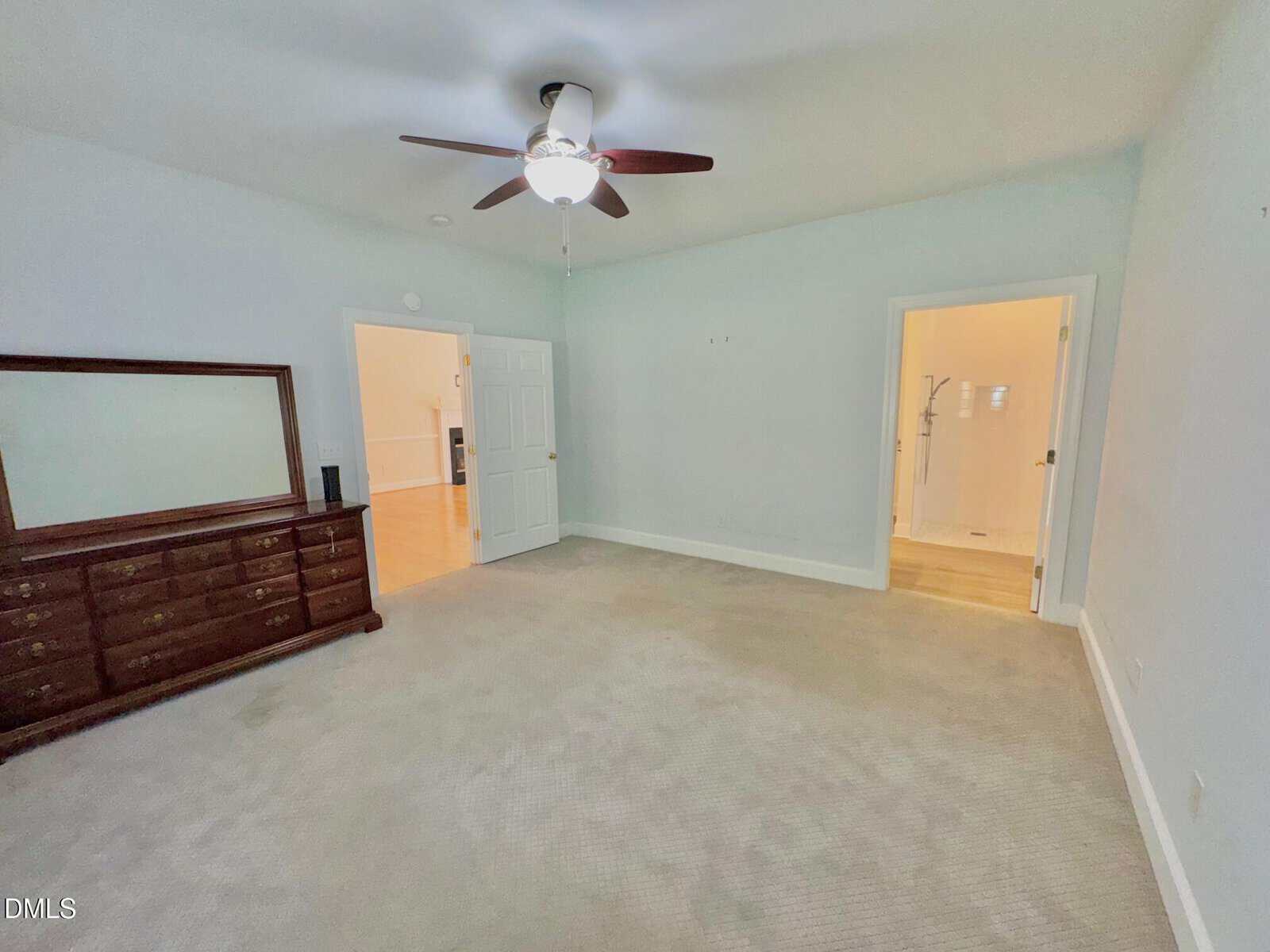 Spacious bedroom with light carpet, ceiling fan, large dresser with mirror, and two doorways leading to other rooms. Walls are light-colored.