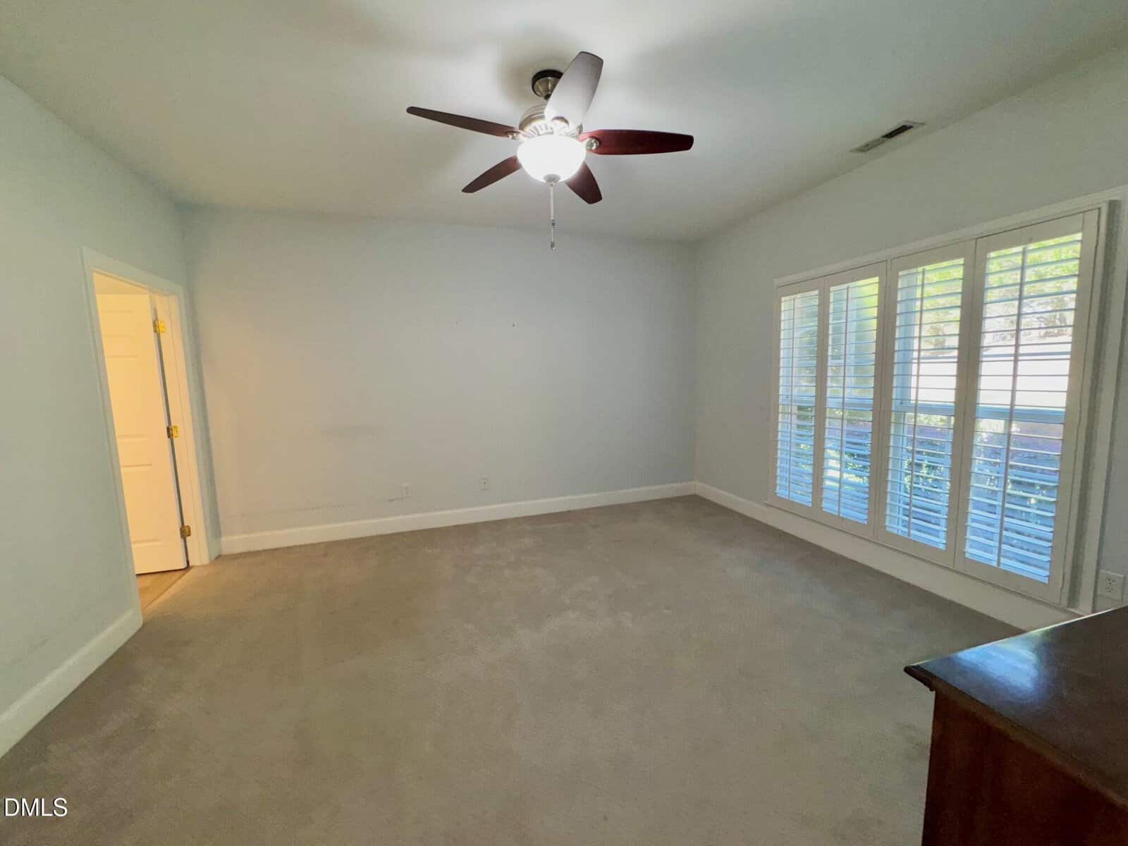 Empty bedroom with beige carpet, a ceiling fan with light, white walls, large window with shutters, and an open door leading to another room. Daylight streams in through the window.