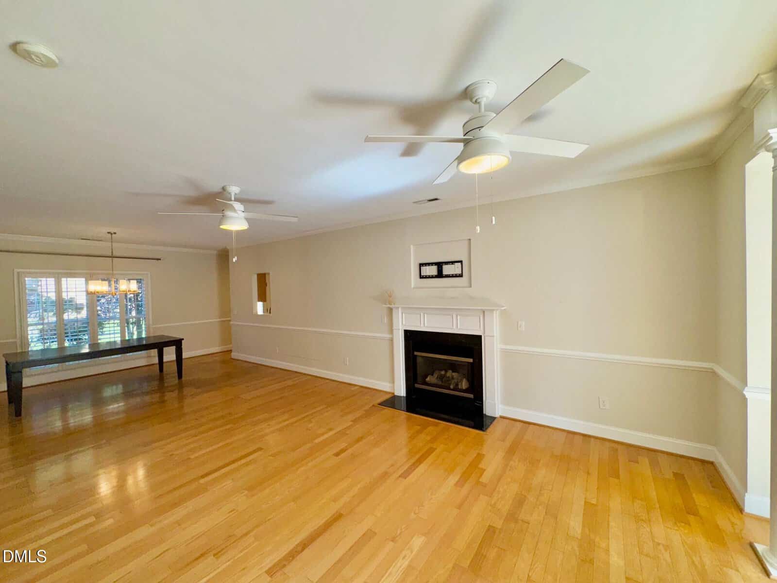 Spacious living room with light wood flooring, a white fireplace centered on the far wall, two white ceiling fans, and a large window providing natural light in the background. A rectangular table is visible on the left.