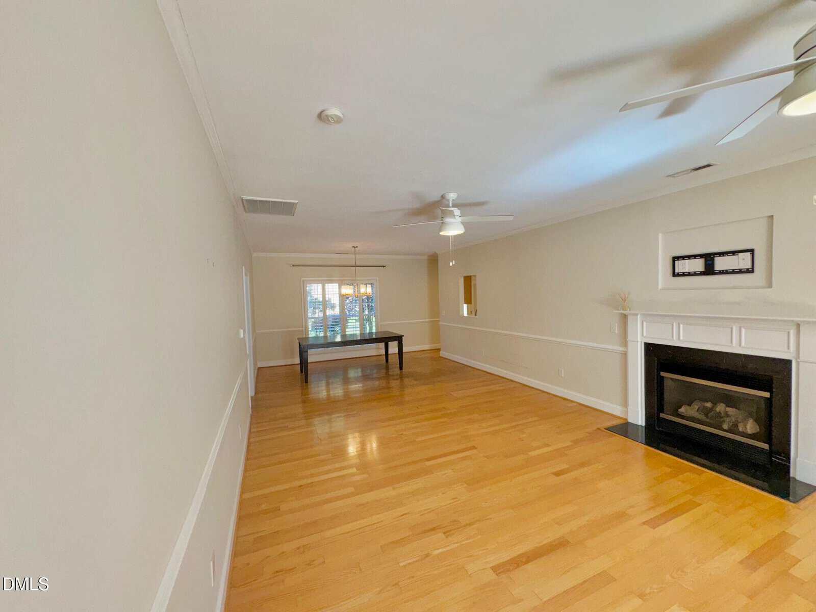 A spacious, empty living room with light wood floors, cream-colored walls, ceiling fans, a fireplace on the right, and a dining table at the far end near two windows with blinds.