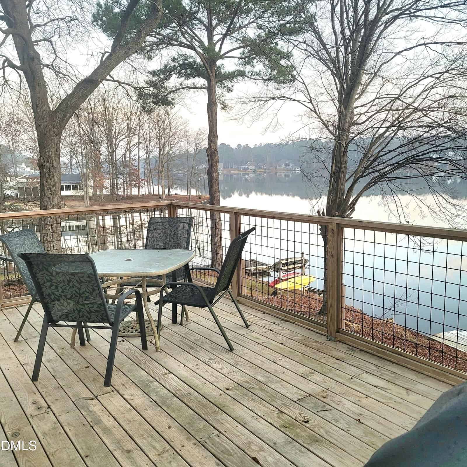 A wooden deck with a glass table and four chairs overlooks a calm lake. Leafless trees surround the area, and a small dock with a yellow kayak is visible by the water. Houses can be seen in the distance.
