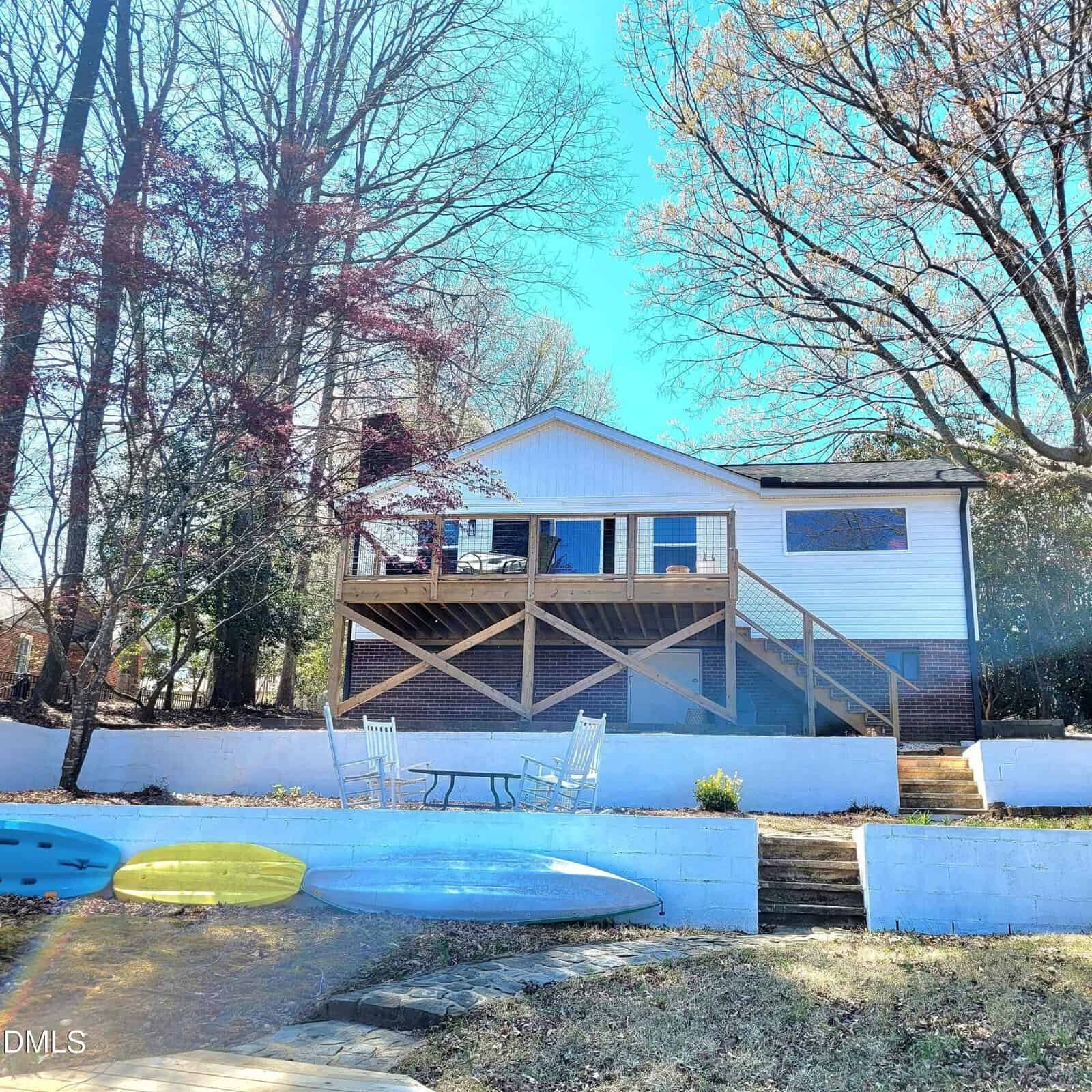 A white house with a raised wooden deck and stairs sits behind a terraced yard at 635 Sagamore Drive in Louisburg, featuring two white chairs and a small table. Two kayaks rest on the grass near a stone path. Leafless trees surround the area under a clear blue sky.