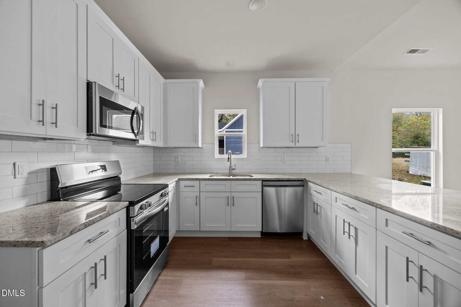 A modern kitchen with white cabinets, stainless steel appliances, granite countertops, white subway tile backsplash, wood flooring, and two small windows above the sink letting in natural light.