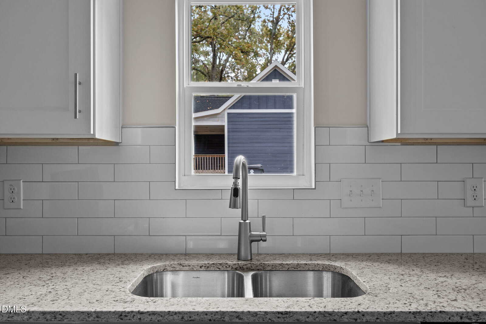 A stainless steel sink with a modern faucet is set in a speckled countertop. White subway tile backsplash and white cabinets frame a window showing a blue house and trees outside.