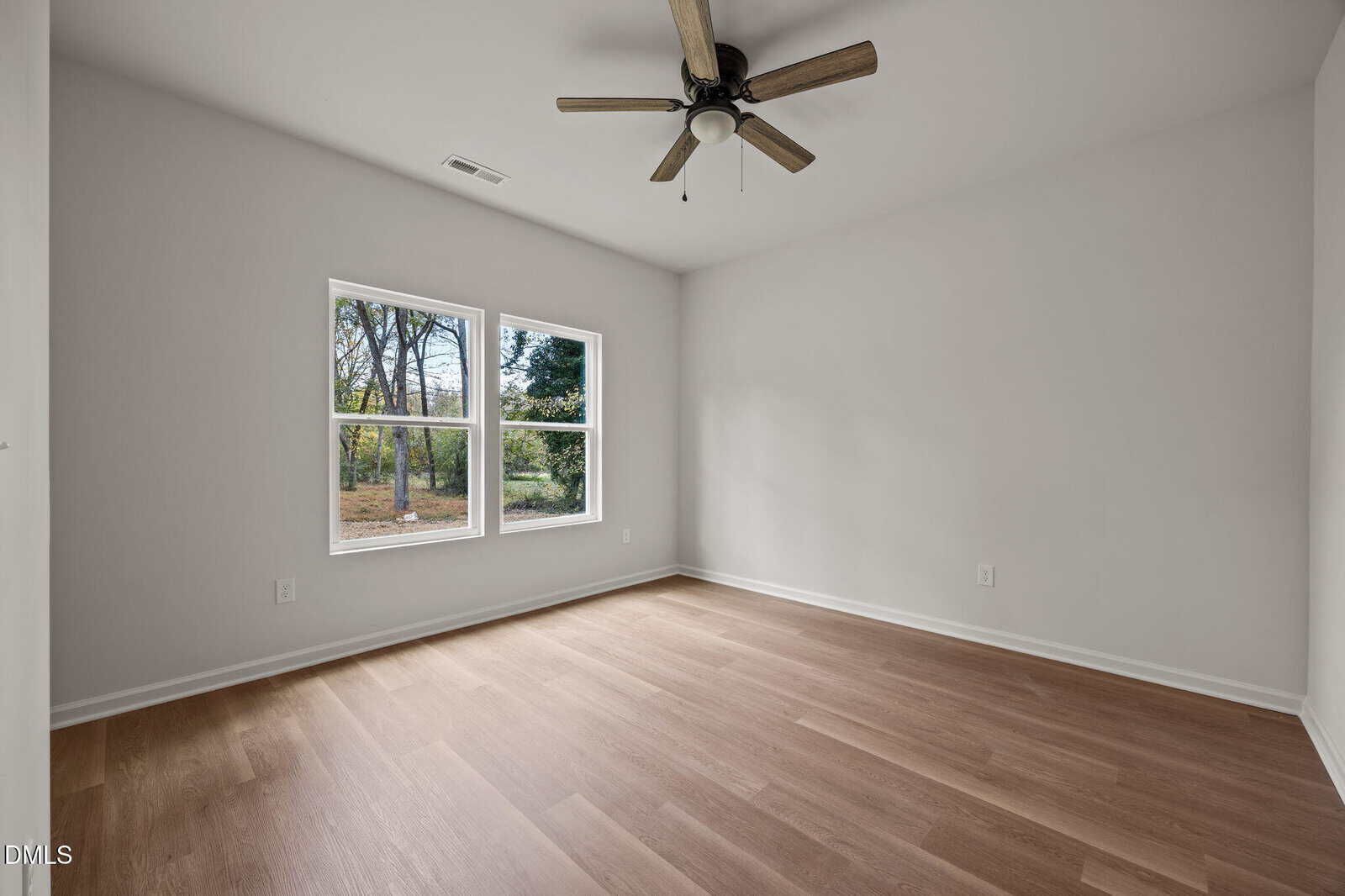 Empty room with light wood floors, white walls, a ceiling fan, and a double window showing trees outside. The room is bright with natural light and has minimal decor.