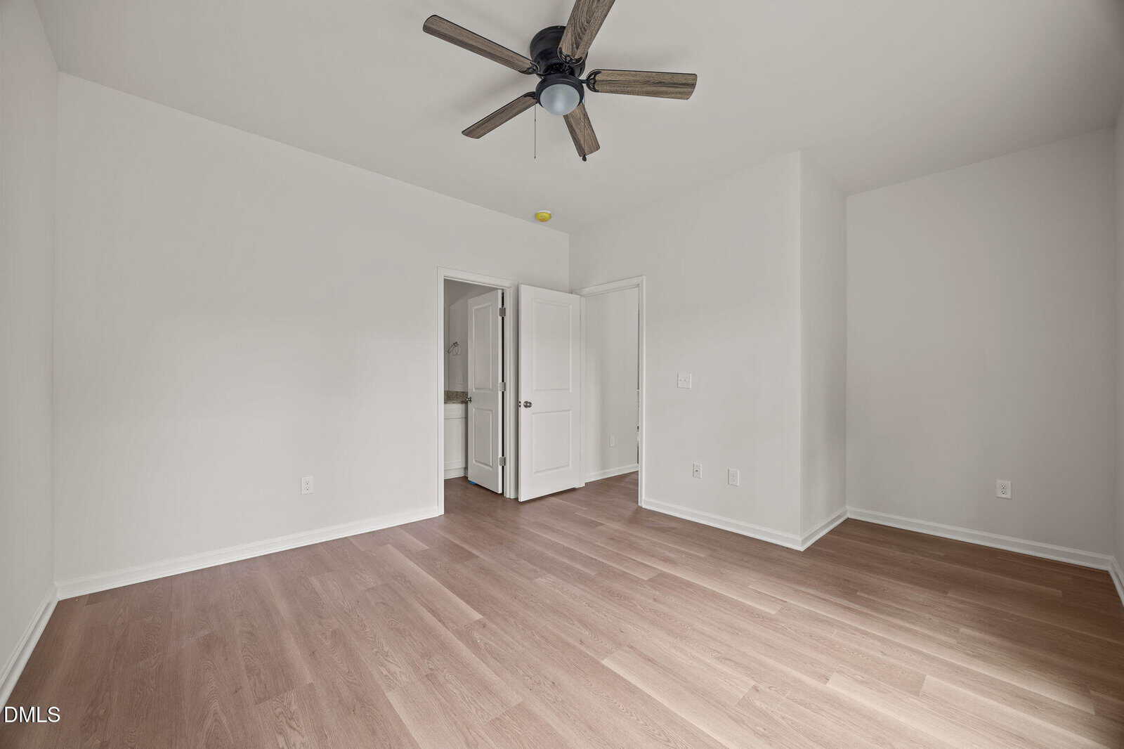 Empty room with light wood flooring, white walls, a ceiling fan, and two open doors—one leading to a bathroom and one to a closet. Natural light brightens the clean, modern space.