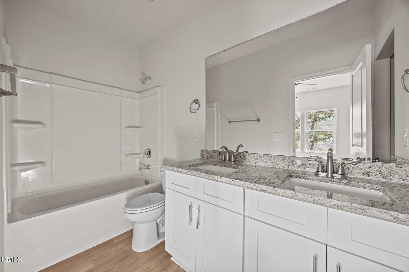 Modern bathroom with double sinks, granite countertop, white cabinets, a bathtub-shower combo, toilet, large mirror, and wood-style flooring. A towel bar and open doorway are also visible.