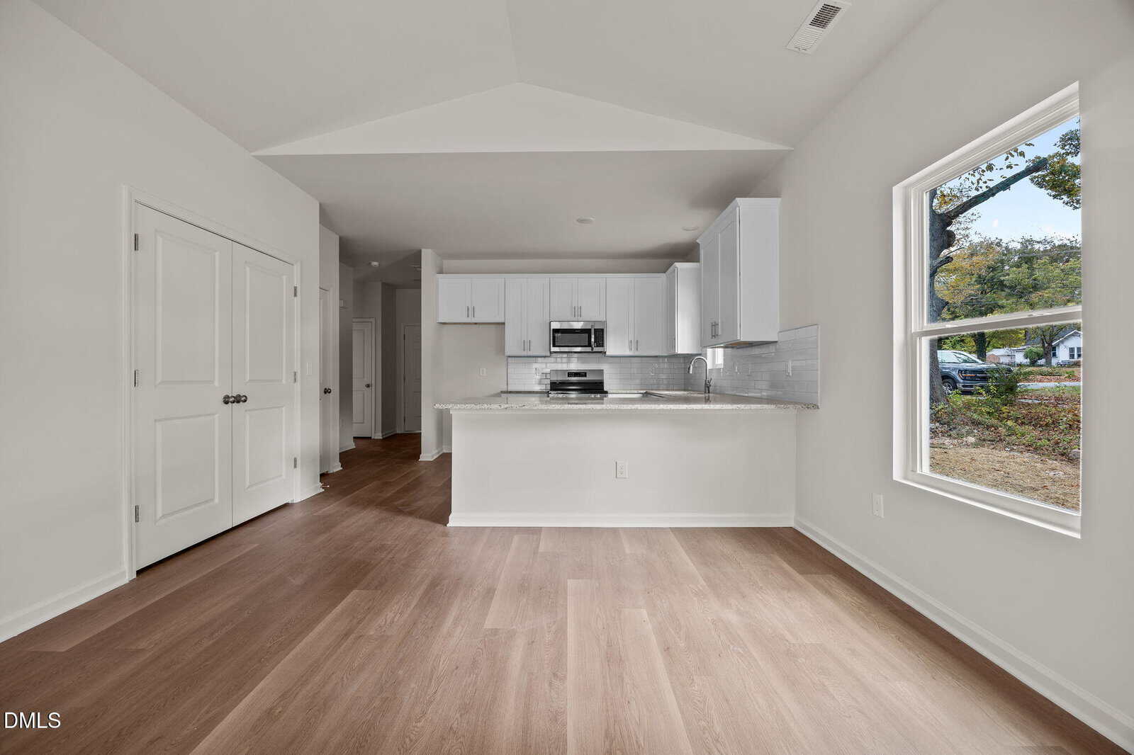 A modern kitchen with white cabinets, stainless steel appliances, and a center island is shown next to an open area with light wood flooring and a large window overlooking a yard with trees.