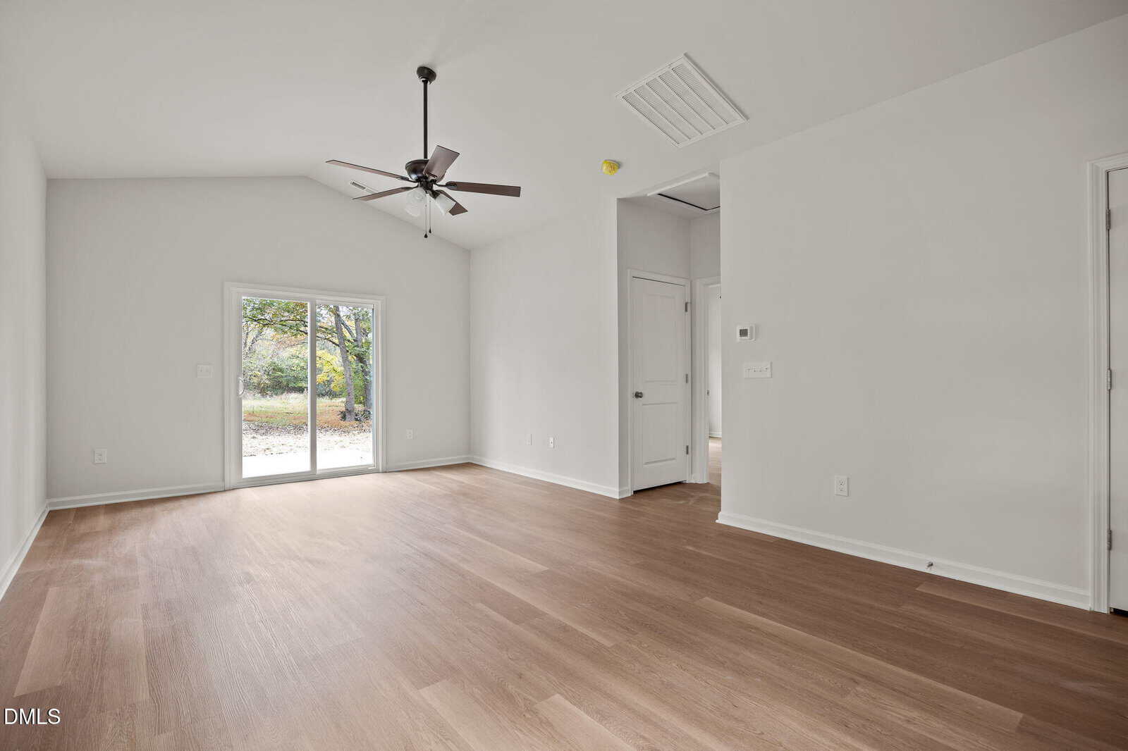 A bright, empty room with light wood flooring, white walls, a ceiling fan, and sliding glass doors leading outside. There are two closed white doors and a small hallway visible on the right.