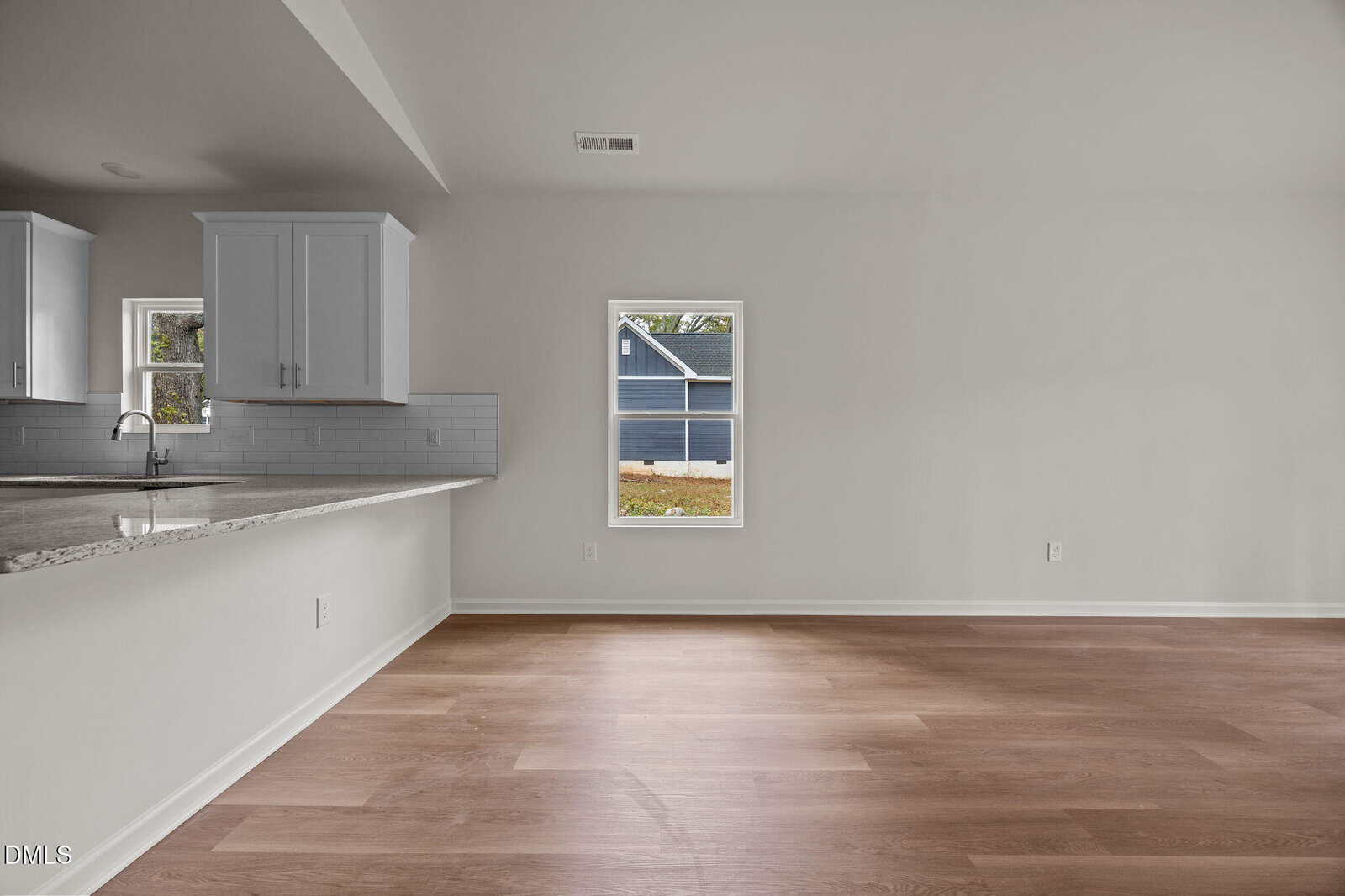 A modern, empty room with light wood flooring, white walls, a window showing part of a blue house outside, and a kitchen area with white cabinets and a granite countertop on the left.