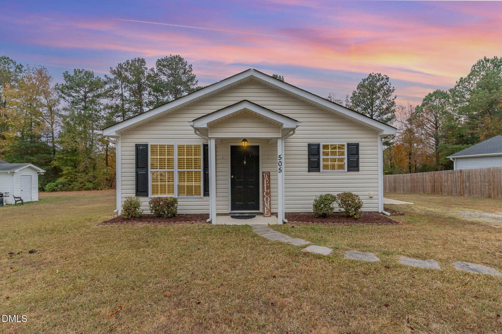 A small, single-story white house with black shutters stands on a grassy yard at sunset on 505 Cotton Avenue in Creedmoor. Stone pavers lead to a black door, two lights, and a vertical welcome sign, with trees lining the background.