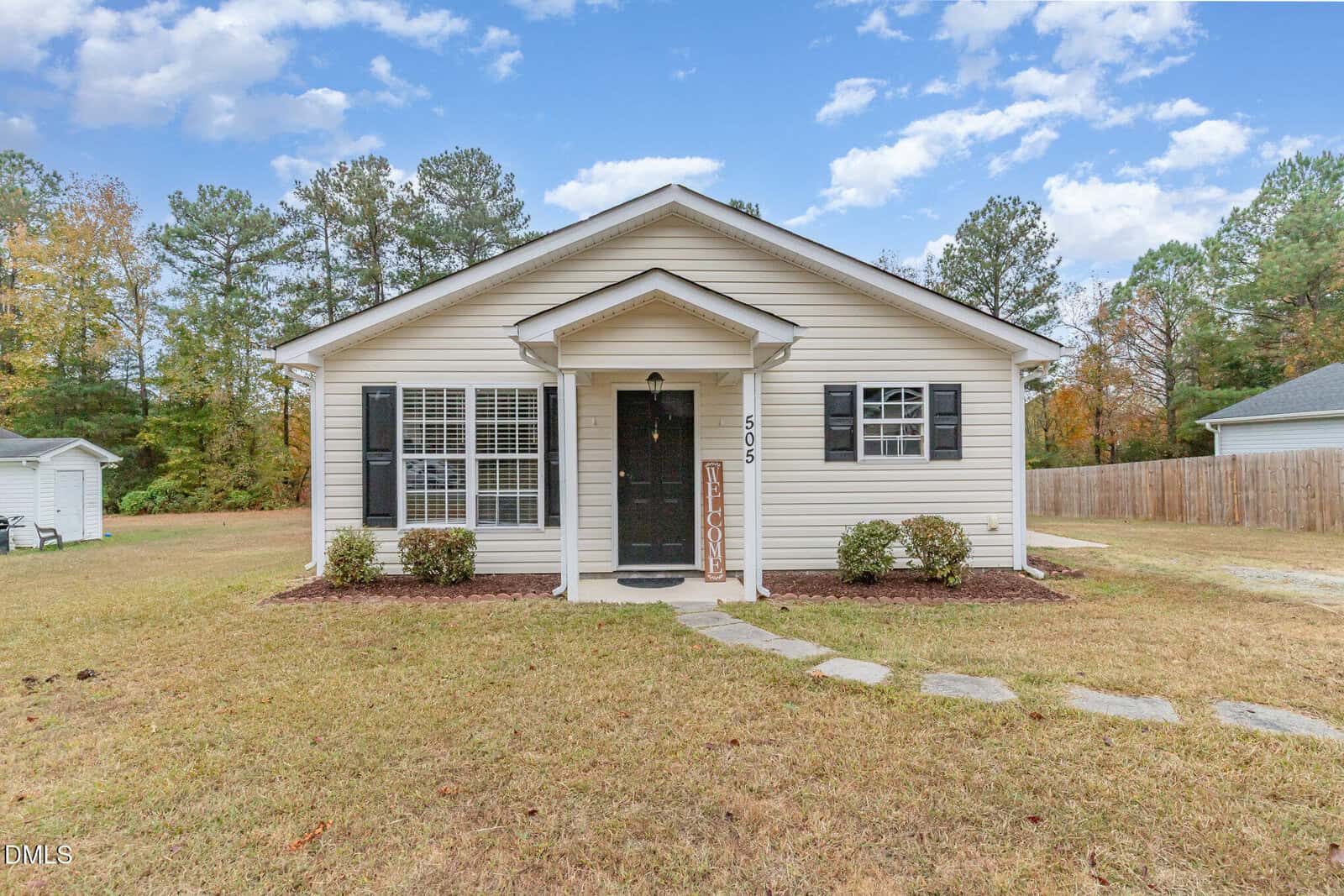 A small, single-story beige house with black shutters and door, front porch with columns, and a stone path leading to the entrance. The lawn is grassy with some bushes and trees in the background.