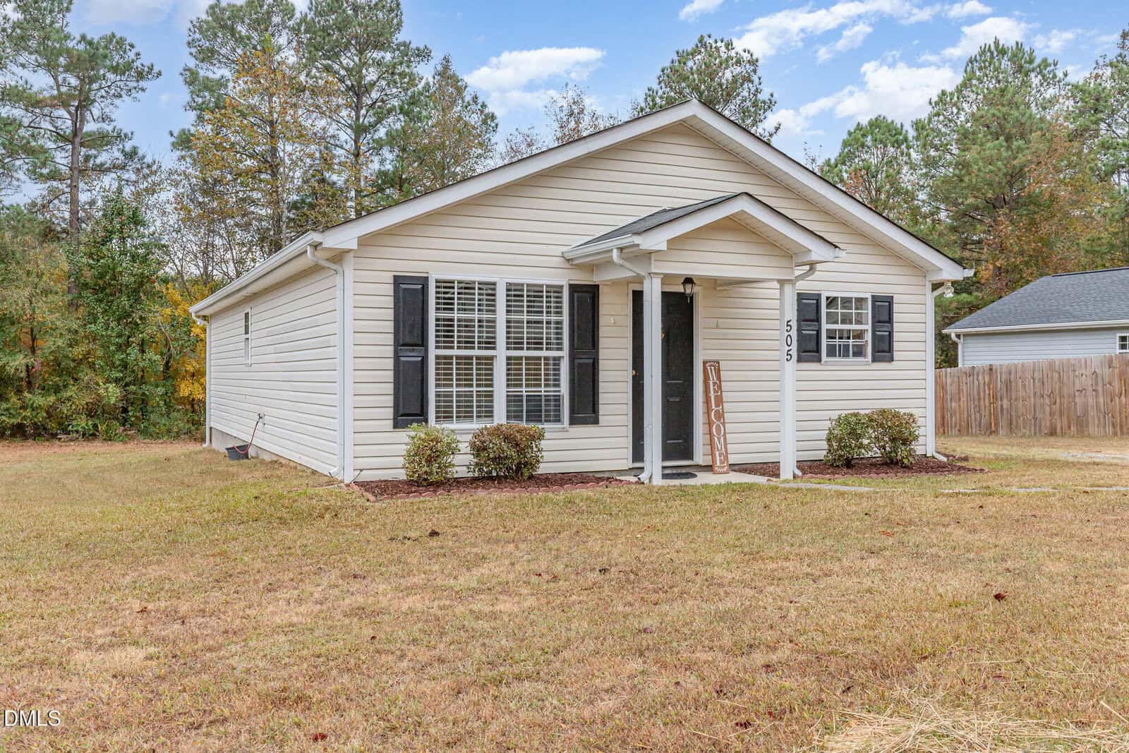 A single-story light-colored house with black shutters and a small front porch, set on a grassy yard with a few shrubs by the entrance and trees in the background.