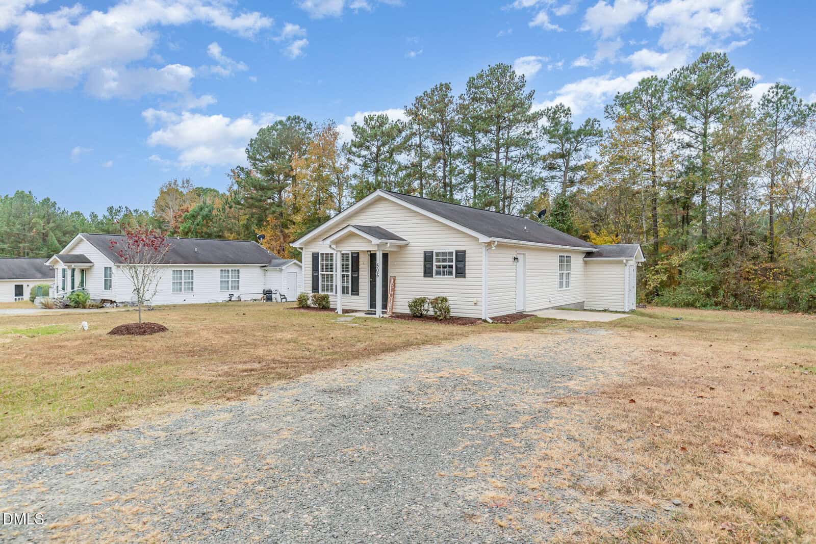 A single-story white house with a covered front porch, set on a large, grassy lot with a gravel driveway. There are trees in the background and another similar house nearby. The sky is partly cloudy.