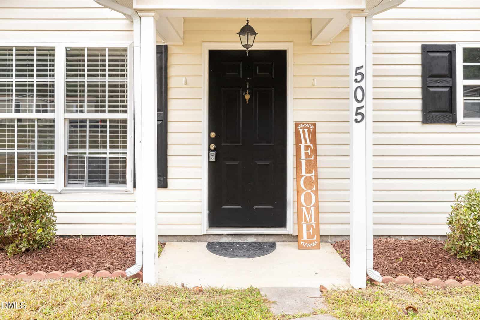 A black front door with a small black lantern above it, beige siding, and white trim. The house number 505 is visible, and a vertical wooden "Welcome" sign is next to the door. Shrubs and windows are on either side.