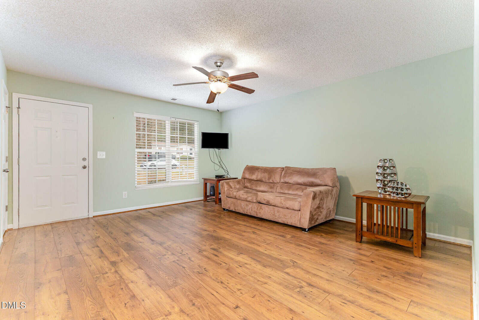 A living room with wood flooring, a beige couch, a small wooden side table with a wine rack, a wall-mounted TV, and a ceiling fan with lights. The walls are light green and there is a window and a white door.