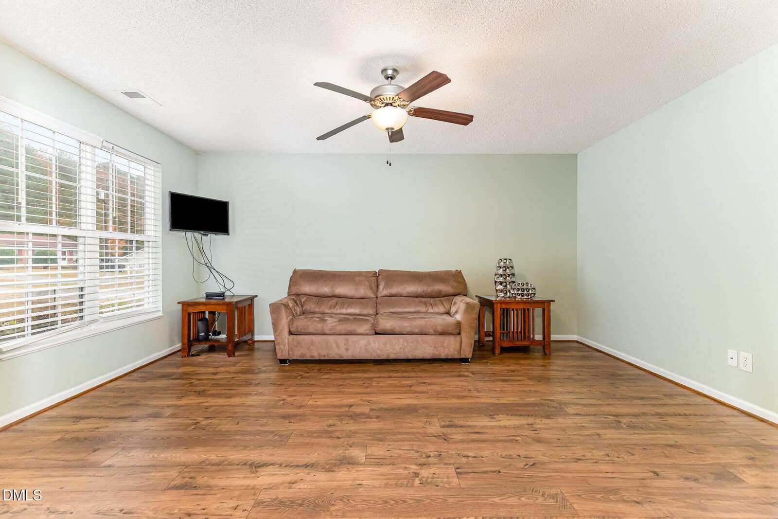 A living room with hardwood floors, a brown sofa against a pale green wall, two wooden side tables, a small wall-mounted TV, and a ceiling fan with lights. Large windows let in natural light from the left.