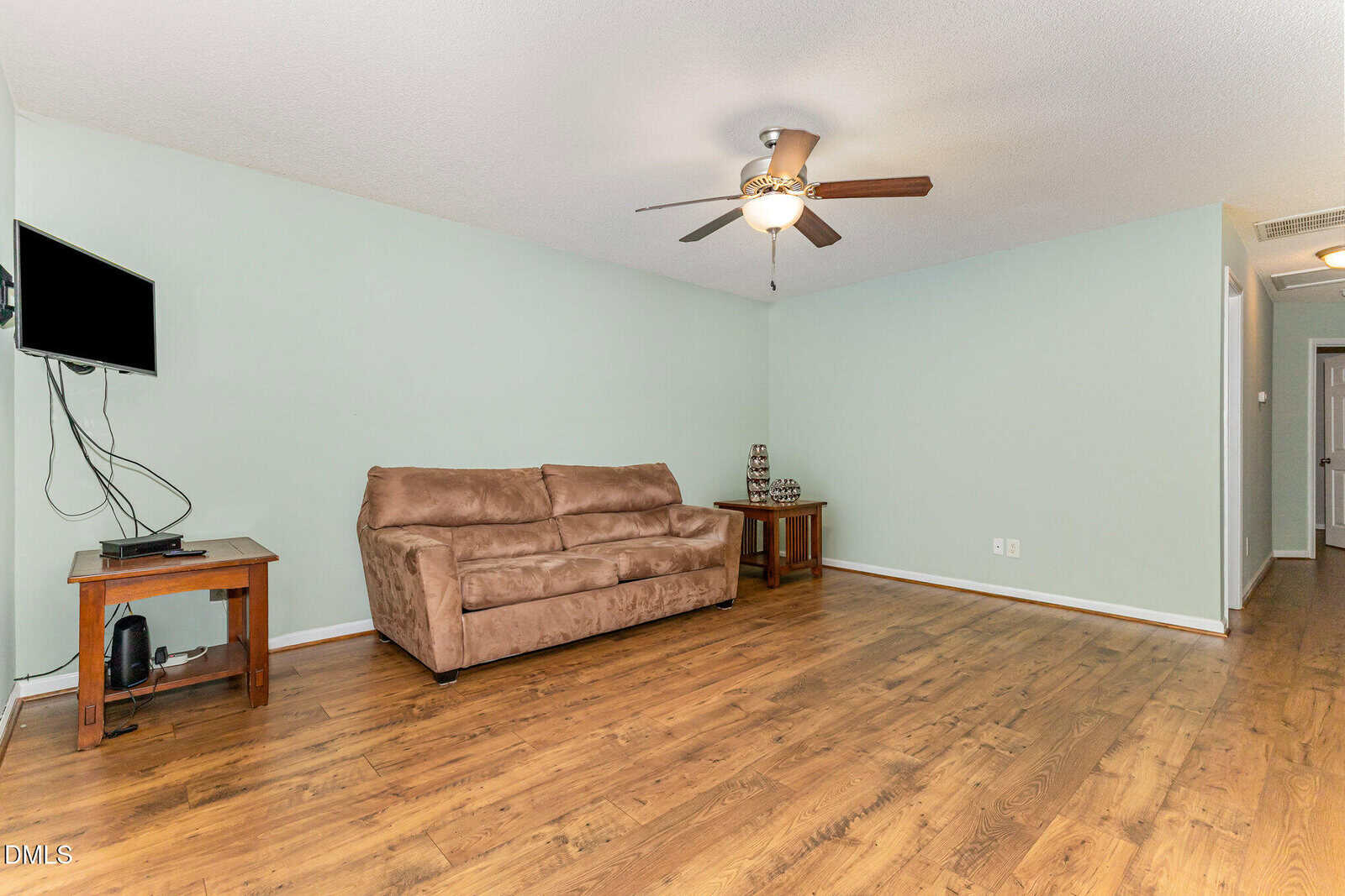 A living room with light green walls, a brown sofa, two wooden side tables, a wall-mounted TV with visible cables, a ceiling fan with light, and wood flooring. A hallway is visible on the right.