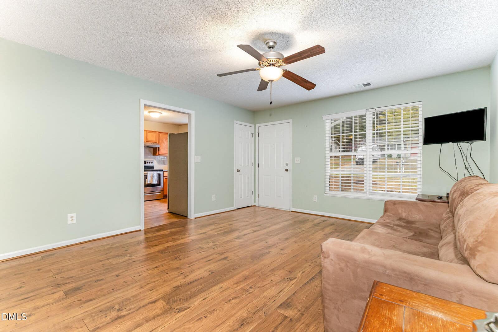 A living room with light green walls, wood flooring, a ceiling fan with lights, a wall-mounted TV, a brown sofa, and a small side table. An open doorway leads to a kitchen with stainless steel appliances.