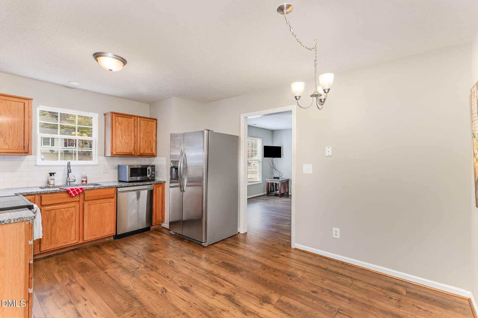 Kitchen with wooden cabinets, stainless steel appliances, granite countertops, and wood flooring. A chandelier hangs above an empty dining area. An open doorway leads to a living room with a TV.