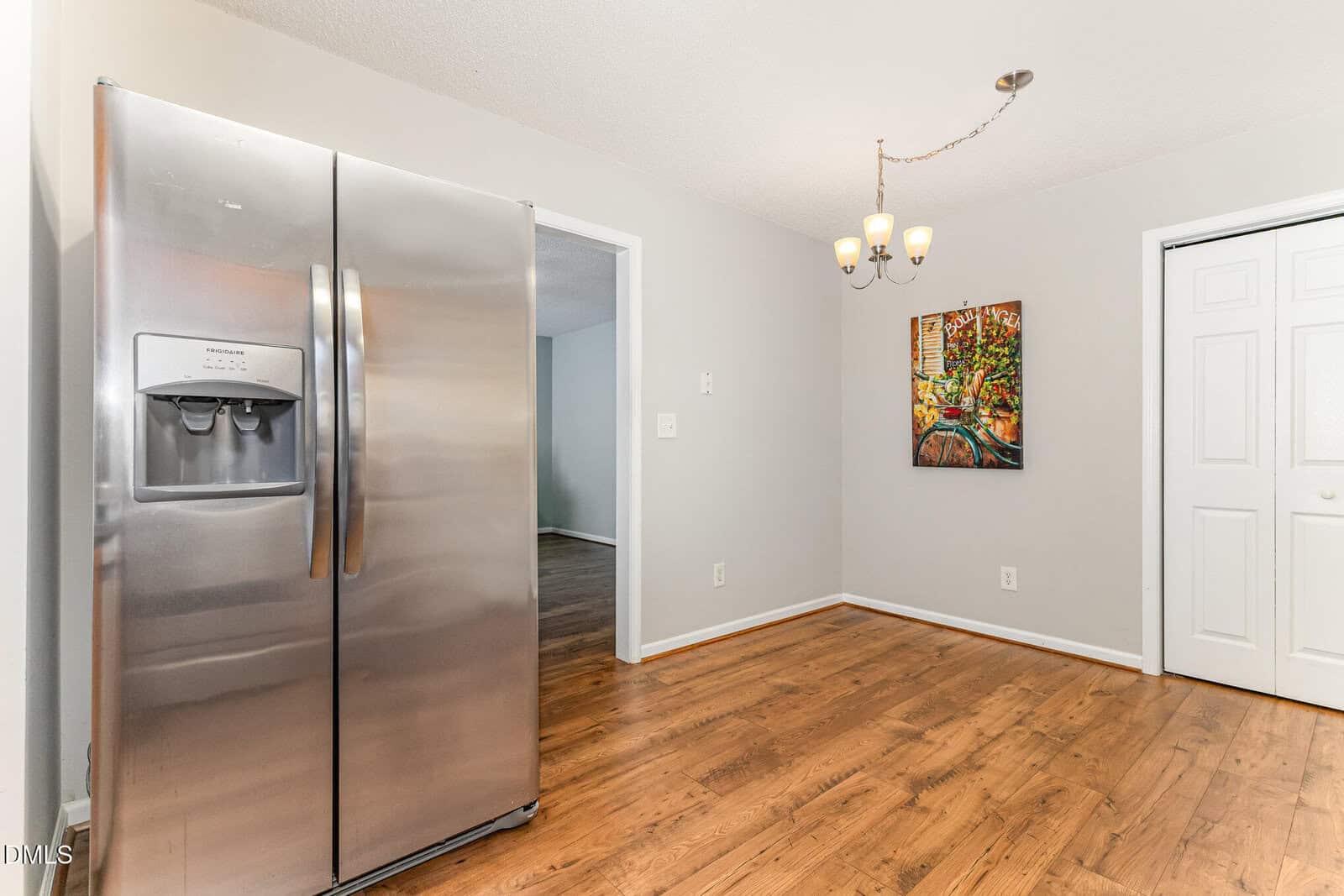 Modern kitchen corner with a stainless steel refrigerator, wooden floors, a white door, a colorful painting on the wall, and a ceiling light fixture with three bulbs.