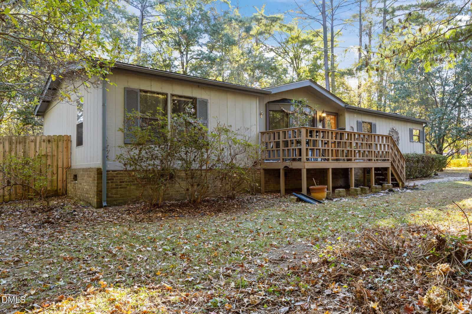 A single-story house with light-colored siding and brick foundation features a wooden front porch with railings, surrounded by trees and fallen leaves on the ground.