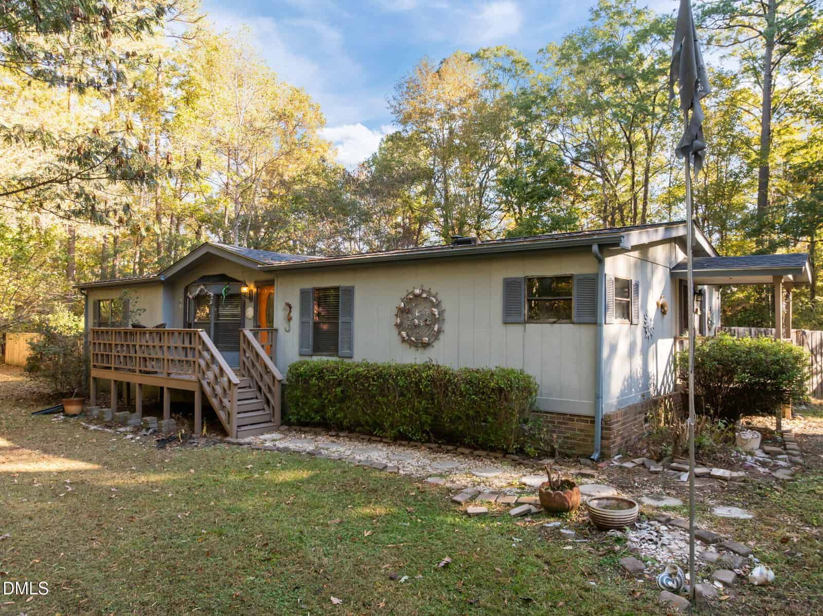 A single-story light blue house with a front porch, a wooden ramp, shuttered windows, and a large decorative piece on the wall. The yard has bushes, a flagpole, and scattered plant pots, surrounded by trees.