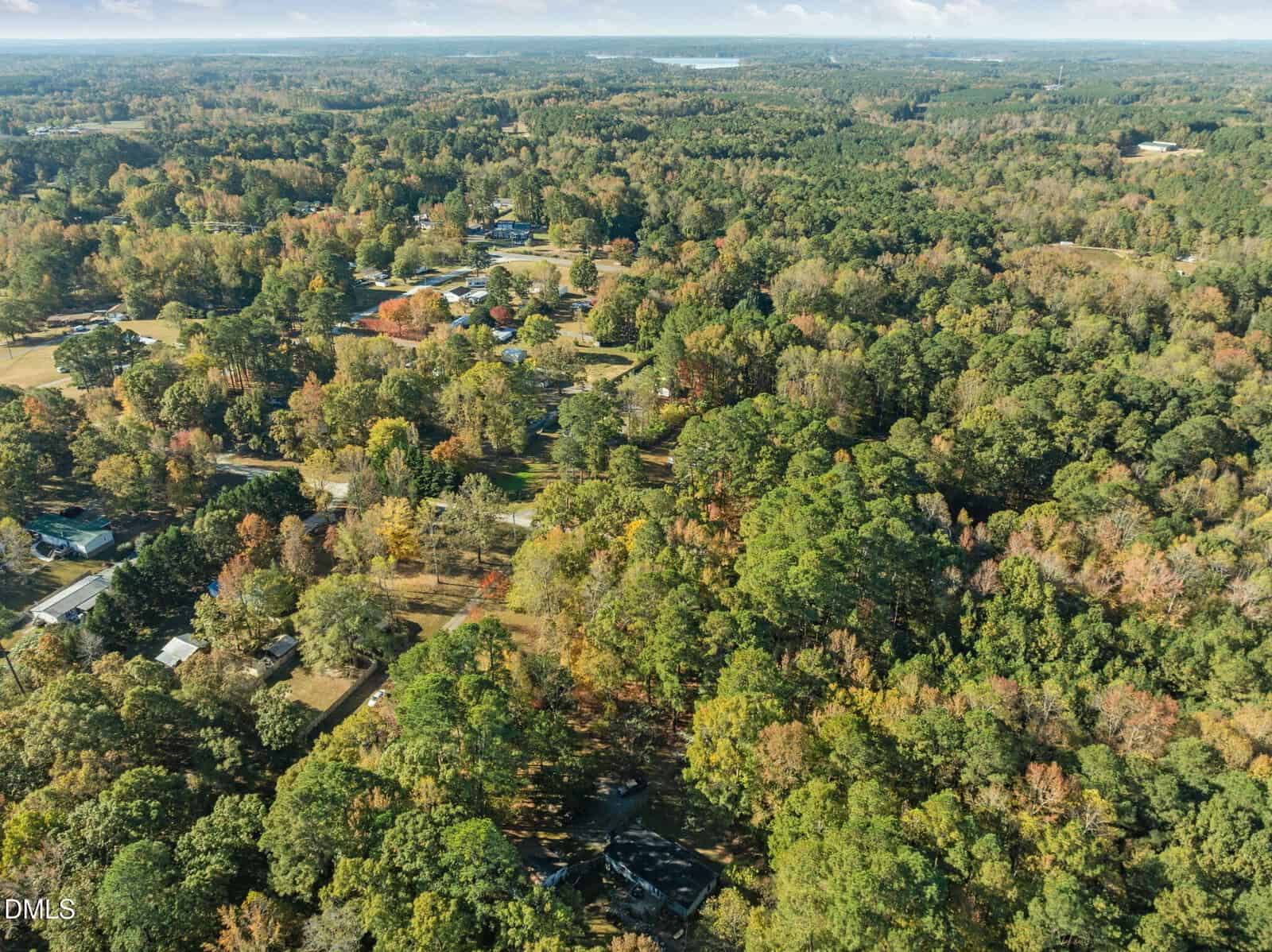 Aerial view of a rural, wooded area with scattered houses and roads surrounded by dense green and autumn-colored trees under a partly cloudy sky.