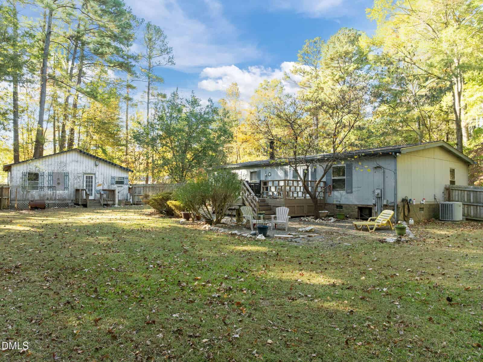 A backyard with grass and scattered leaves, featuring a small fire pit area with chairs, two light-colored single-story buildings, trees in the background, and a partly cloudy sky overhead.