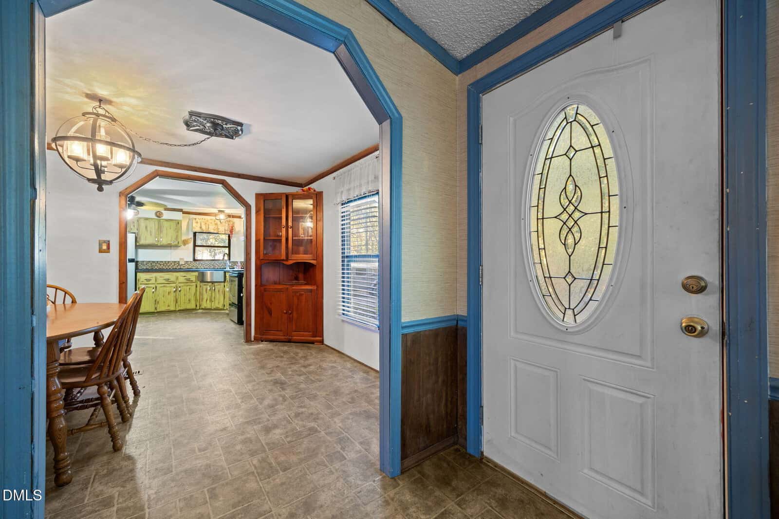 A view from the entryway shows a white door with an oval window, a dining area with a wooden table and chairs, a large light fixture, and a kitchen with green cabinets in the background.