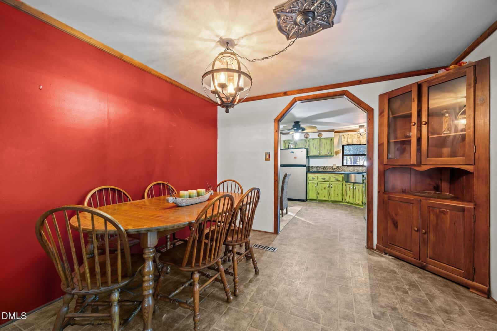Dining area with a wooden table and six chairs next to a red accent wall, rustic wooden corner cabinet, and view into a kitchen with green cabinets and white appliances.