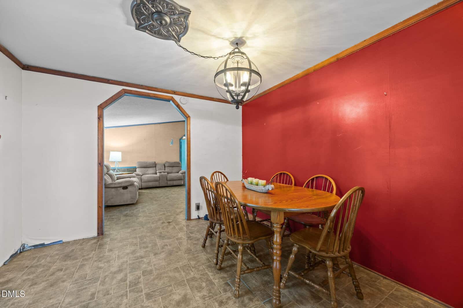 A dining area with a wooden table and five chairs sits beside a bold red accent wall. An ornate ceiling light fixture hangs above. An arched doorway leads to a living room with beige sofas in the background.