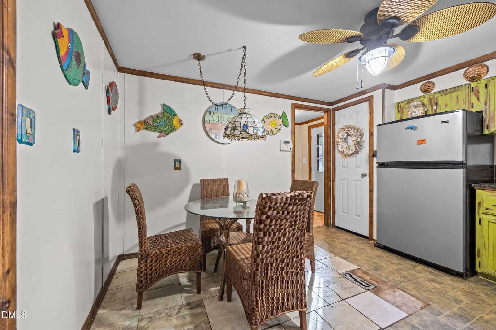 A cozy dining area with a glass table, four wicker chairs, fish-themed wall decor, ceiling fan, and a kitchen with a silver fridge and lime-green cabinets in the background.