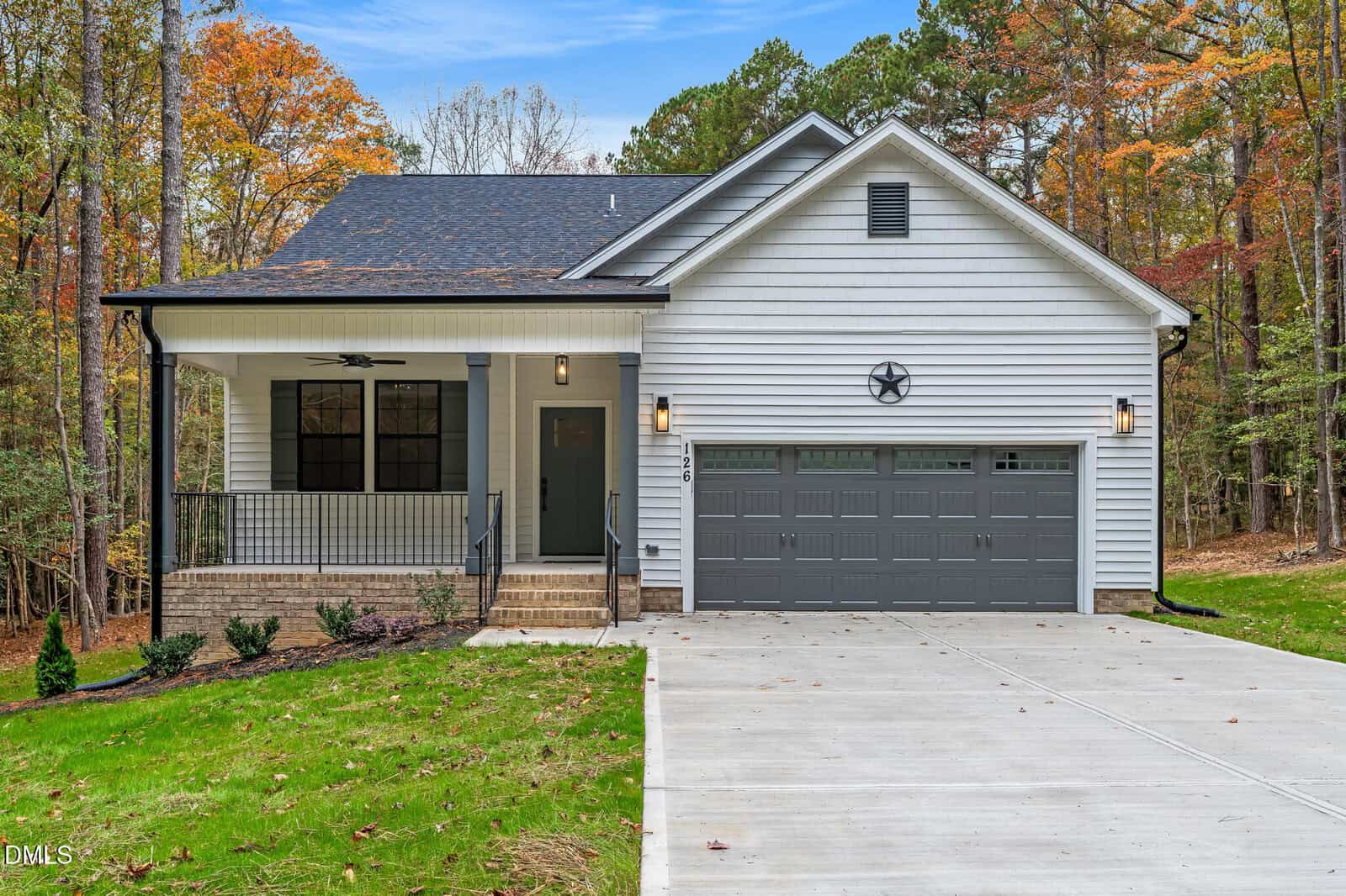 A modern single-story house at 126 Acoma Drive in Louisburg features crisp white siding, a two-car gray garage, a welcoming covered front porch with steps leading to the entrance, and is surrounded by a wooded yard adorned with vibrant autumn trees. A charming star decorates the front exterior.